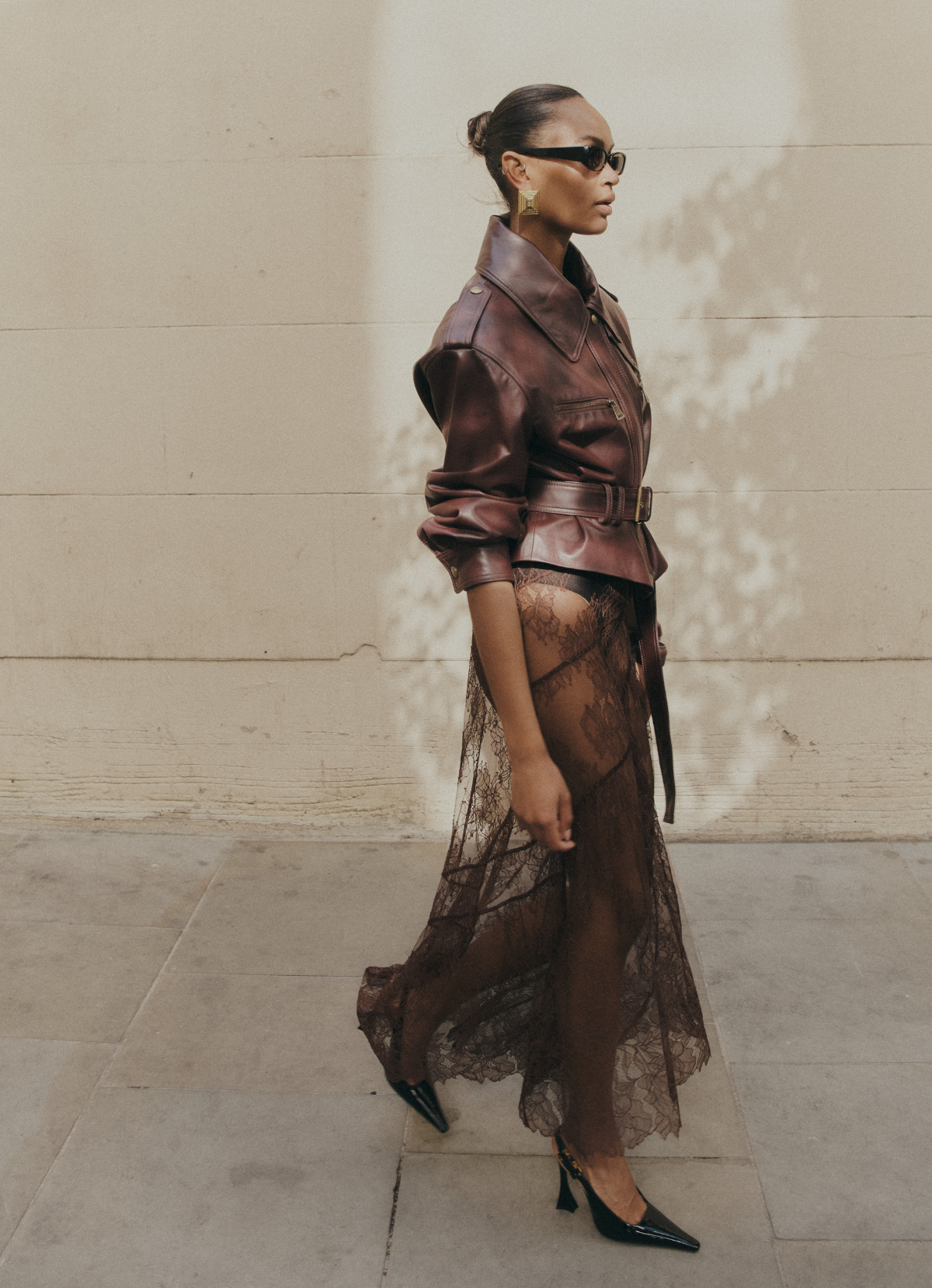 Image of model walking wearing burgundy cropped leather jacket with sleeves pushed up and jacket fastened with belt.