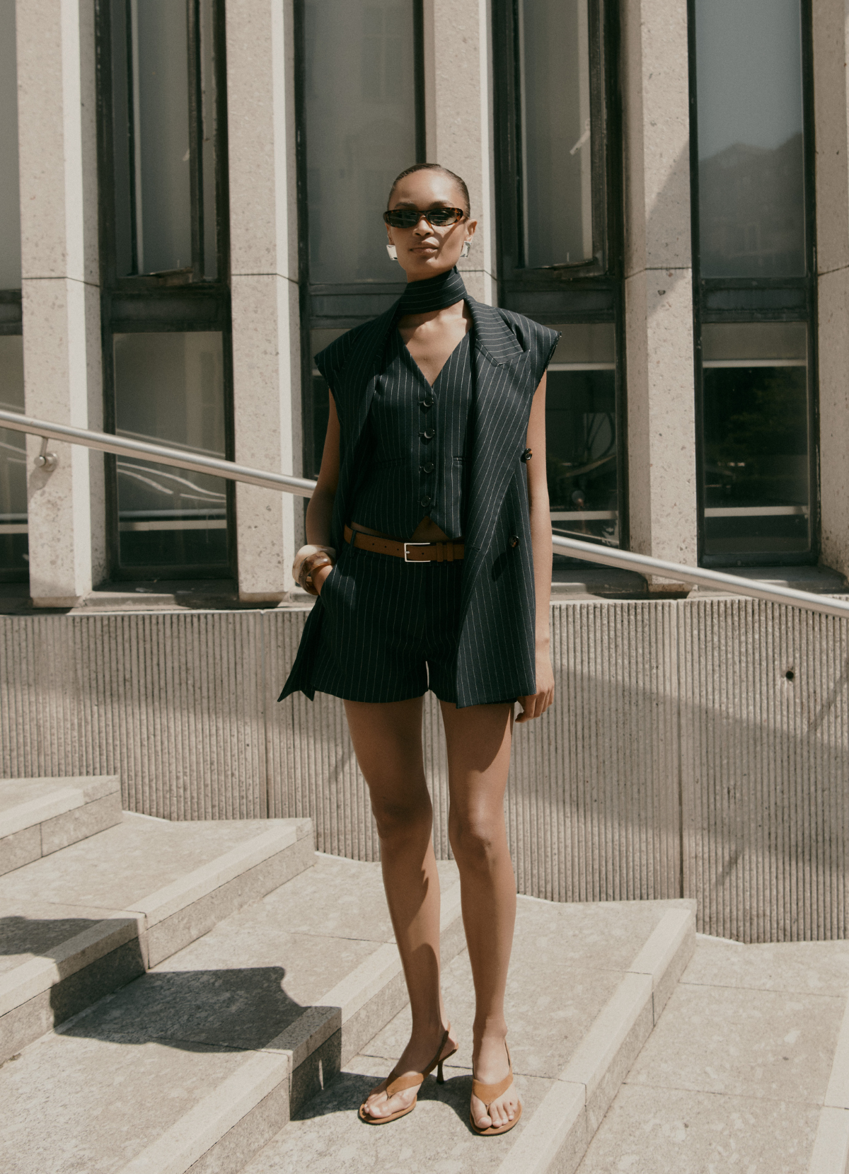 Photo of model standing in outdoor city environment wearing navy pinstripe sleeveless blazer and shorts with one hand in pocket.